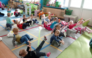 przedszkolaki siedzą na dywanie, ręcei nogi uniesione do góry
