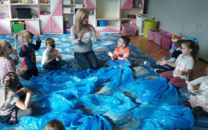 dzieci siedzą wkole,dłonie uniesione do góry, na środku niebieska folia malarska
