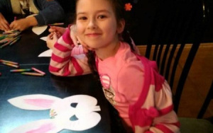 uśmiechnięta dziewczynka siedzi przy stole nad papierową głową kruliczka
