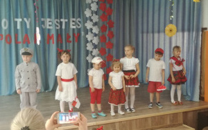 Dzieci w strojach biało czerwonych na scenie. W tle napis Kto Ty jesteś? Polak Mały!