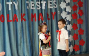 dziewczynka w stroju krakowskim i chłopiec z czerwoną kokardą pod kołnierzykiem mówią do mikrofonu
