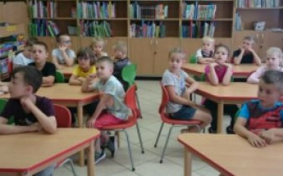 dzieci w czytelni siedzą przy stolikach,w tle regały z książkami
