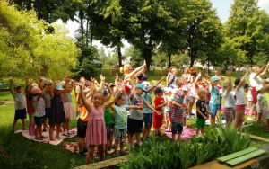 Przedszkolaki stoją na trawie, ramiona uniesione do góry, śpiewają hymn przedszkola