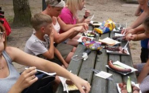 dzieci i rodzice zajadają kiełbaski przy drewnianych stołach