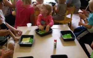 Dzieci i rodzice przy stolikach, na nich klej i papierowe elementy w pudełkach