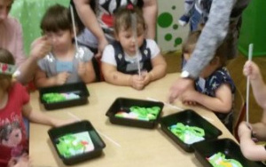 Dzieci przy stolikach, trzymają w rączkach patyczki