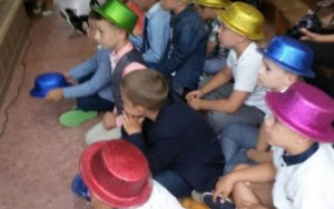 Dzieci w kolorowych, brokatowych kapeluszach siedzą pod sceną.