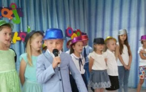 Chłopiec w niebieskim,brokatowym kapeluszu mówi domikrofonu, w tle grupa dzieci