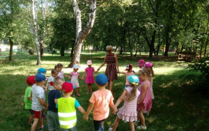 Dzieci między drzewami,na zielonej trawie bawią się w kole ze swoją panią