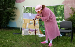 Aktorka w przebraniu babci w fioletowym, puchowym szlafroku, czepku i okularach