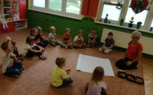 zainteresowane dzieci  siedzą na dywanie, ciocia Ania objaśnia czekające ich zadanie, przed nią arkusz papieru,flamastry i kredki