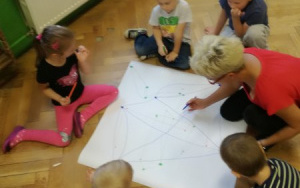 piątka dzieci i ciocia Ania pochyleni nad arkuszem papieru, zaznaczają mazakami kolorowe punkty i kreślą linie