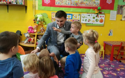 Pan Patryk trzyma chłopca na kolanach i pokazuje ilustracje w książce,obok inne dzieci