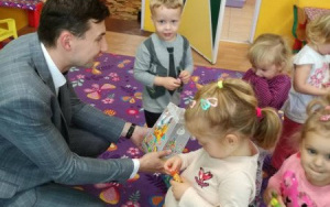 Pan Patryk z książkąw ręceprzykuca na dywanie,uśmiecha się do dzieci z cukierkami w dłoniach