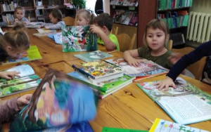 dzieci siedząprzy drewnianych stołach, oglądają kolorowe książki