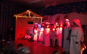 Dzieci w jasełkowych strojach zbierają się do występu przy drewnianej szopce,na scenie