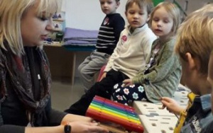 kobieta w szarym swetrze trzyma kolorowy instrument podobny do cymbałków, chłopiec uderza w niego pałeczką