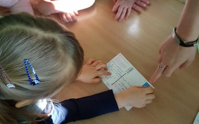 dziewczynka przykleja znaczek pocztowy na pocztówce w miejscu wskazanym przez nauczycielkę