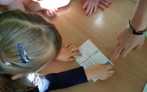 dziewczynka przykleja znaczek pocztowy na pocztówce w miejscu wskazanym przez nauczycielkę