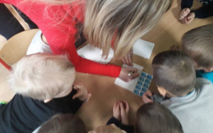 Pani Martynka wraz z dziećmi nachylona nad stolikiem, wypisuje pocztówkę, nad pocztówką leżą znaczki pocztowe