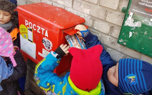 Dziecko w czerwonej czapce i kolorowej kurtce wrzuca pocztówkę do czerwonej skrzynki na listy