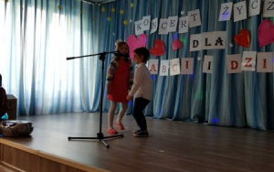 dziewczynka w czerwonej sukience i chłopiec w białej koszuli tańczą przy mikrofonie i śpiewają piosenkę