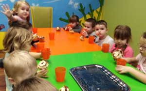 dzieci przy zielonym i pomarańczowym stole zajadają słodkie bułki i piją sok z pomarańczowych kubków, na stole srebrna taca