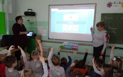 dzieci siedzą przed tablicą interaktywną, sygnalizują podnosząc do góry rękę, chęć rozwiązania wyświetlanego na tablicy zadania