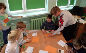 dzieci przy stoliku sklejają puzzle z kartek pocztowych  z pomocą pani nauczycielki