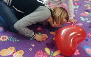 kobieta dmucha w czerwony balon położony na dywanie, obok przygląda się temu dziewczynka z różową kokardą we włosach