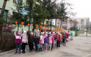 dzieci stoją przed budynkiem przedszkola, do góry unosząkolorowe,papierowe kwiaty
