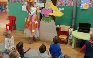 Nauczycielka w stroju Paniu Wiosny pokazuje dzieciom papierową sylwetę Wiosny,której suknię trzeba ozdobić kwiatami