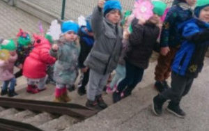 Dzieci w wiosnennych kurtkach z papierowymi kwiatami w dłoniach wchodzą po schodach na osiedlu