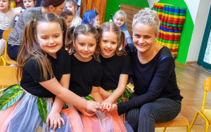 trzy dziewczynki w czarnych koszulkach i tiulowych,kolorowych spódniczkach siedzą na ławce zadowolone ze swojego występu, razem z uśmiechniętą ciocią Anią trzymają się za ręce