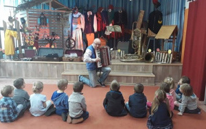 mężczyzna przyklęka na jednymkolanie, prezentuje zainteresowanym dzieciom akordeon, w tle wystawa regionalna
