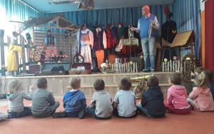 Mężczyzna na scenie prezentuje dzieciom instrumenty dęte. Dzieci siedzą na dywanie, w tle wystawa regionalna