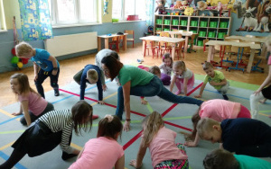 dzieci na dywanie próbują wspólnie z panią Kornelią wykonać ćwiczenie sportowe - jedna noga w wykroku do tyłu, druga ugięta w kolanie z przodu