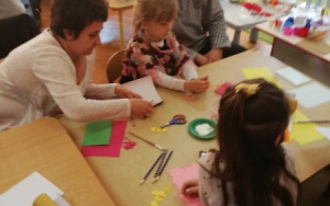 dzieci z rodzicami przy stolikach przyklejają wycięte elementy do brystolowych kartek