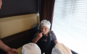 dziewczynka w kucharskiej czapce degustuje z białej łyżeczki ciepłą czekoladę