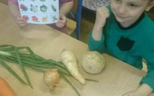 dwoje dzieci przy stoliku, dziewczynka trzyma kartę pracy z ilustracjami warzyw, przed nimi: szczypior, cebula, marchew,pietruszka i seler