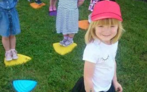 uśmiechnięte dzieci w czerwonych czapeczkach stoją na kolorowych,plastikowych kamieniach rozłożonych w trawie
