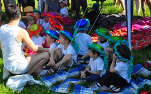dzieci w kolorowych,brokatowych kapeluszach przewiązanych bibułową szarfą siedzą na kocu rozłożonym na trawie