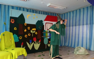 scena, na niej aktorka - zielona wróżka. w tle dekoracja - kwiecista łąka, drzewa i domek