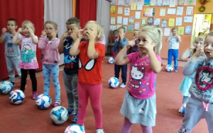 skupione 5. latki stoją z paluszkami na oczach, przed nimi piłki do piłki nożnej