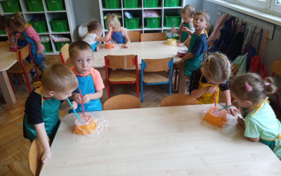 dzieci w kolorowych fartuszkach stoją przy stolikach, dmuchają słomkami w wodę z mydłem- robią bańki