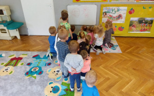dzieci siedzą na dywanie, wspólnie z ciocią rysują na tablicy brzydkie bakterie