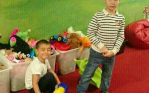 dwóch chłopców na czerwonym dywanie, een przykuca przy ciężarwce, drugi stoi, w tle pudła z zabawkami oraz malunek kolejki