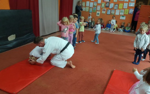 dzieci ustawione w dwuszeregu na dywanie przed dwoma czerwonymi materacami. Przy ednym z nich przykuca mężczyzna w białym kimonie, pomaga dziecku zrobić przewrót w przód