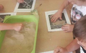 Dzieci smarują obrazek klejem i posypują go piaskiem