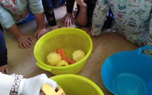 dzieci zgromadzone przy stole na którym stoją: zielona i niebieska miska oraz sitko z umytymi marchewkami i jabłkami. Dłonie w jednorazowych rękawiczkach ścierają na tarce do miski, marchewkę
